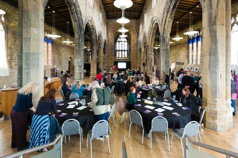 Groups of people sitting around several table looking towards a speaker in the distance. This is all in a converted church. 