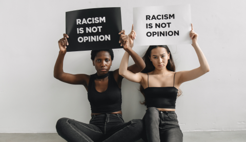 Two women sitting on the ground, next to each other and facing the camera, holding signs above their heads that read, "Racism is not opinion".